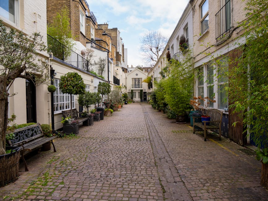 A narrow, cobblestone courtyard surrounded by white and beige townhouse-style buildings with black iron balconies and window frames. The courtyard is lined with potted plants, small trees, and shrubs